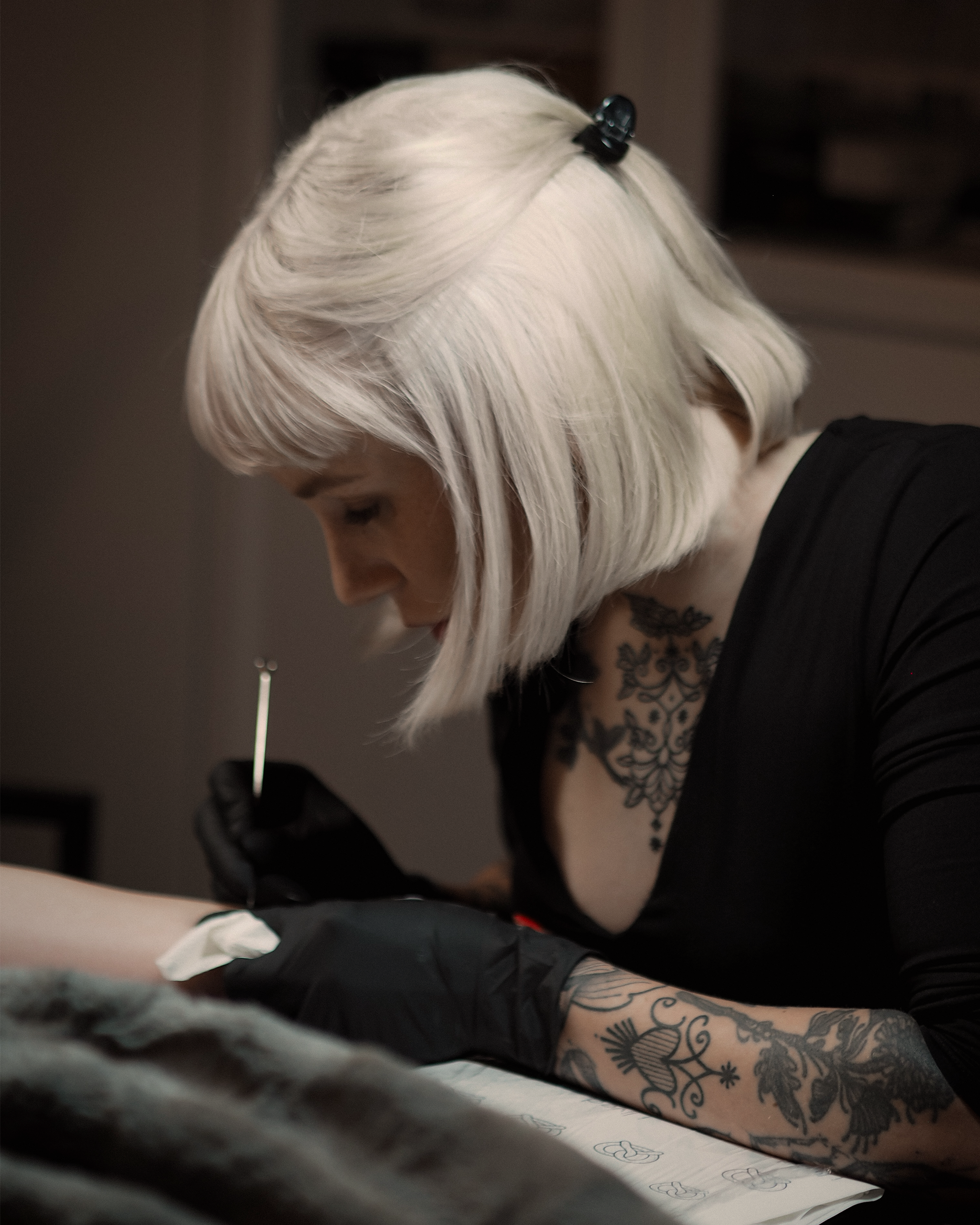 Man sieht Ramona Hozzel, die Gründerin von Heart and Poke, die Speziallisten für Sommersprossen / Freckles und Handpoke Tattoos. Sie hat Blonde Haare und ist gerade am Stechen am Tattoo. Sie trägt ein schwarzes Top und ist im Profil zu sehen.