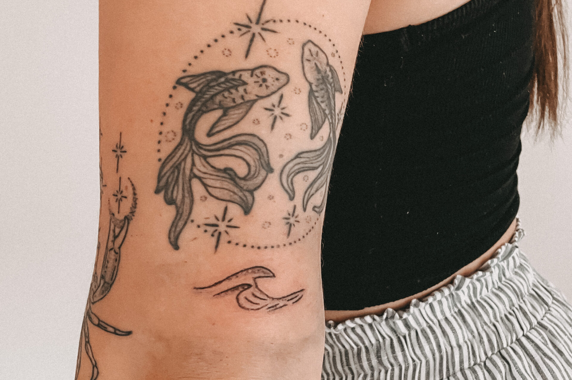 Diese Bild wird für Sommersprossen und Handpoke Tattoos von Heart and Poke verwendet und man sieht eine Nahaufnahme von einem Frauenoberarm auf dem ein Tattoo mit zwei Fischen zu sehen ist.