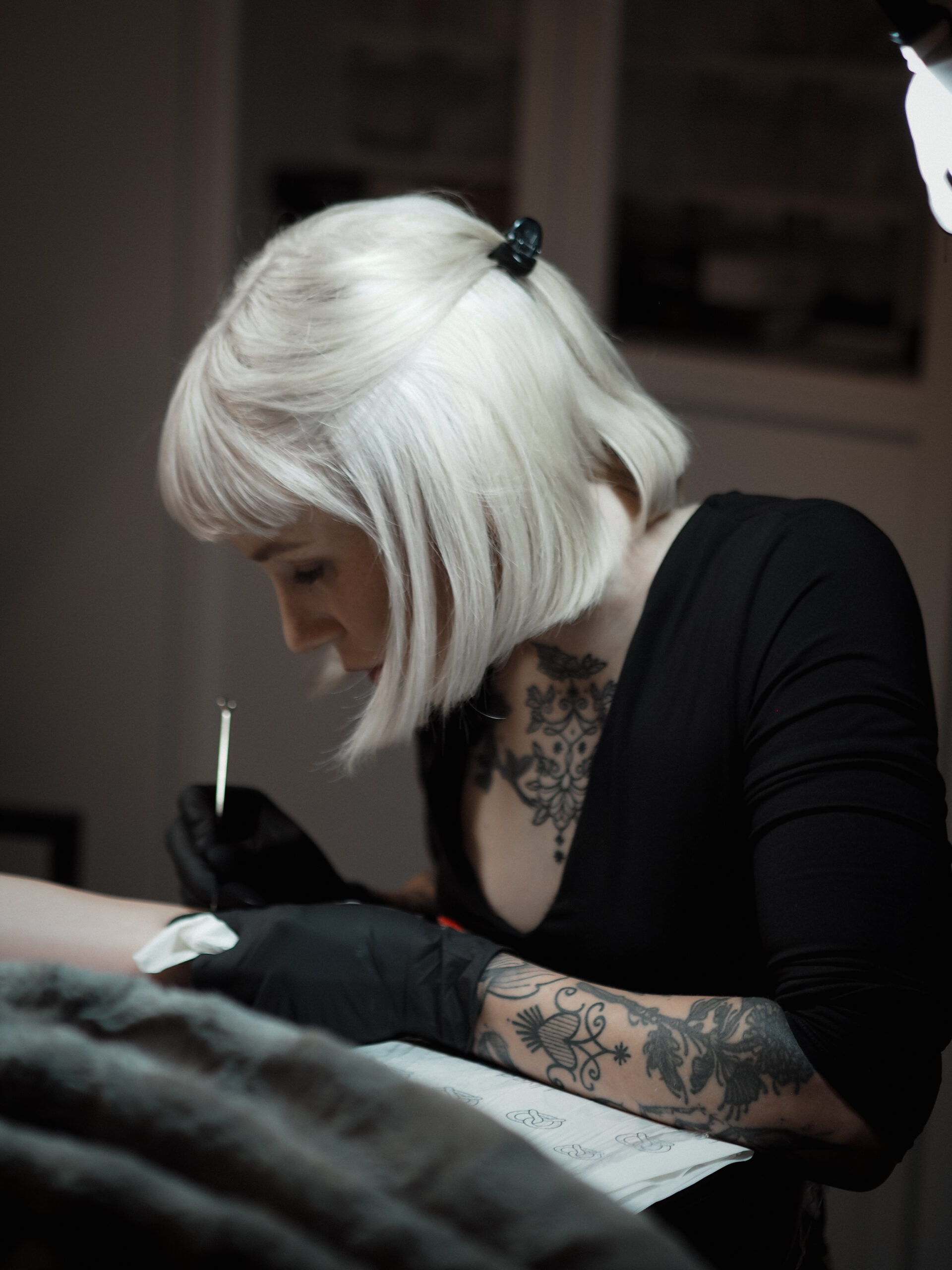 Man sieht Ramona Hozzel, die Gründerin von Heart and Poke, die Speziallisten für Sommersprossen / Freckles und Handpoke Tattoos. Sie hat Blonde Haare und ist gerade am Stechen am Tattoo. Sie trägt ein schwarzes Top und ist im Profil zu sehen.
