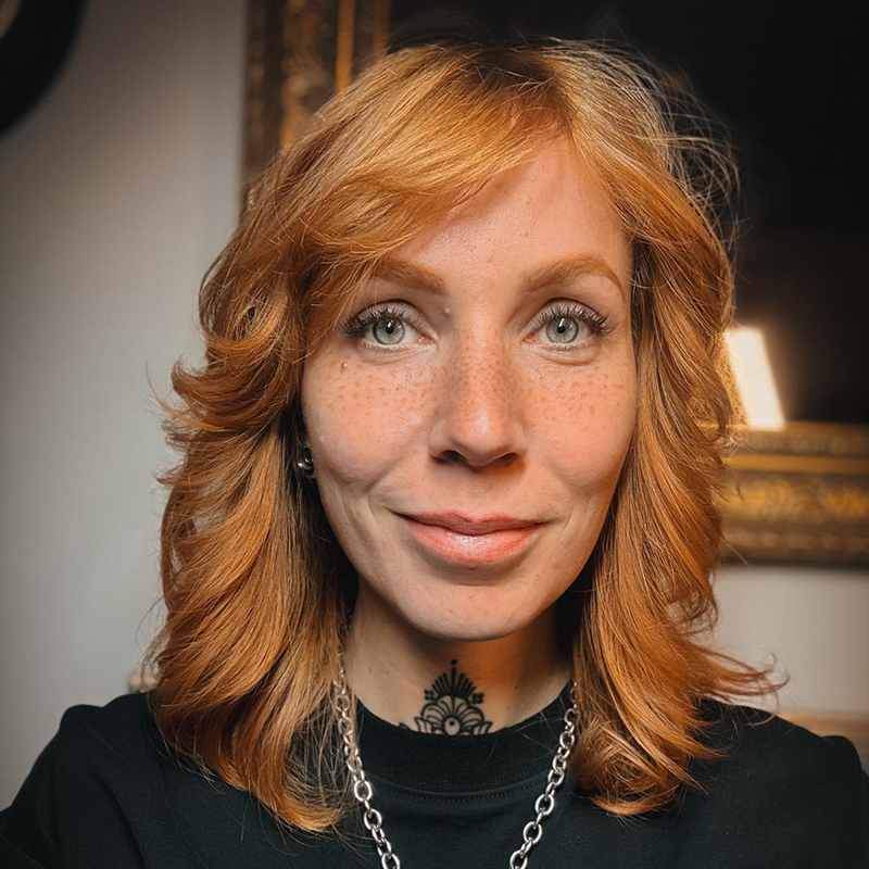 Ein frontales Foto von Mona Hozzel der Geschäftsführerin von Heart and Poke. Sie trägt ein schwarzes Oberteil und hat einen Ponny schnitt mit Orangen Haaren.